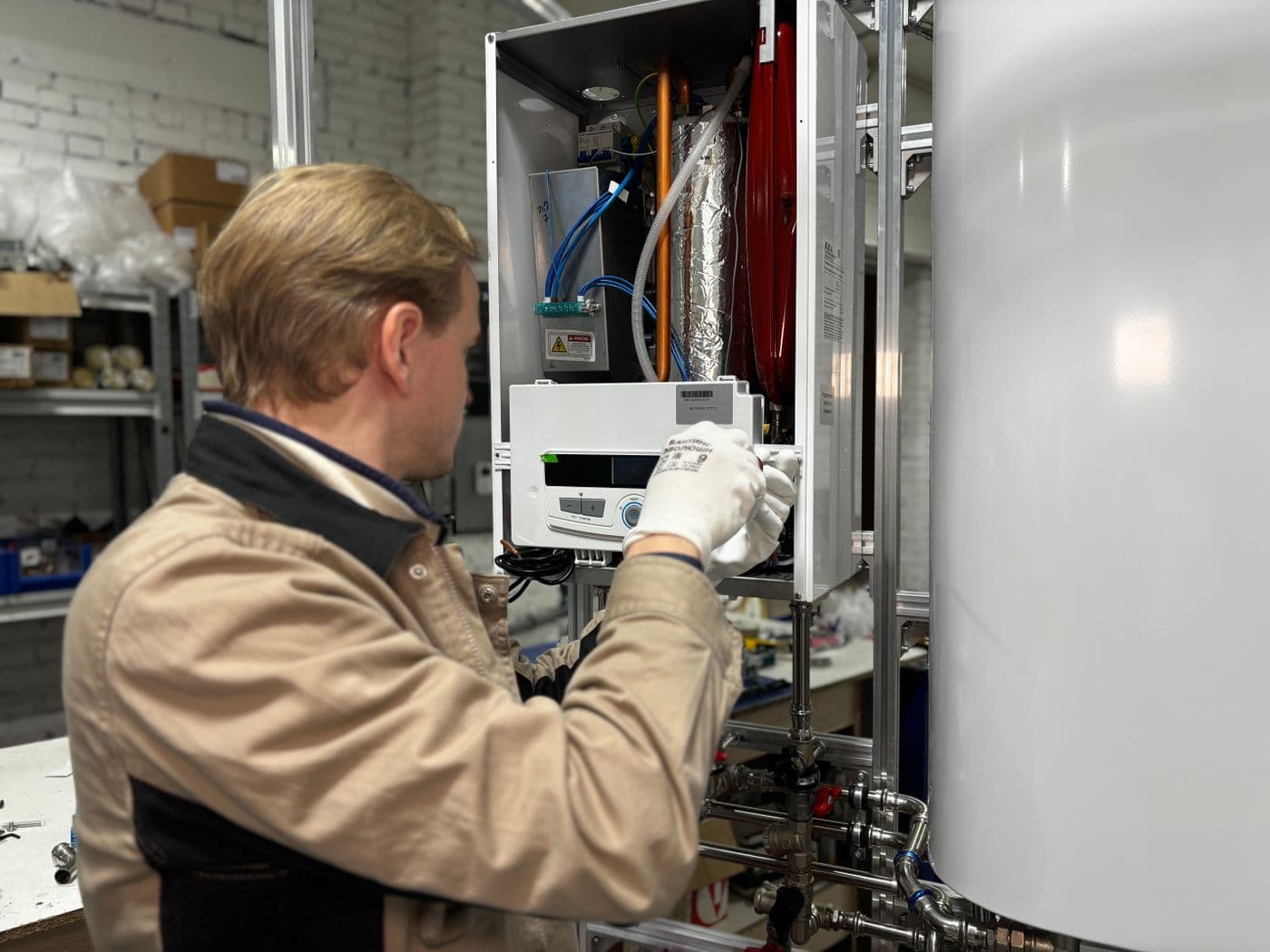 A heating engineer working inside an open wall-mounted boiler next to a hot water cylinder