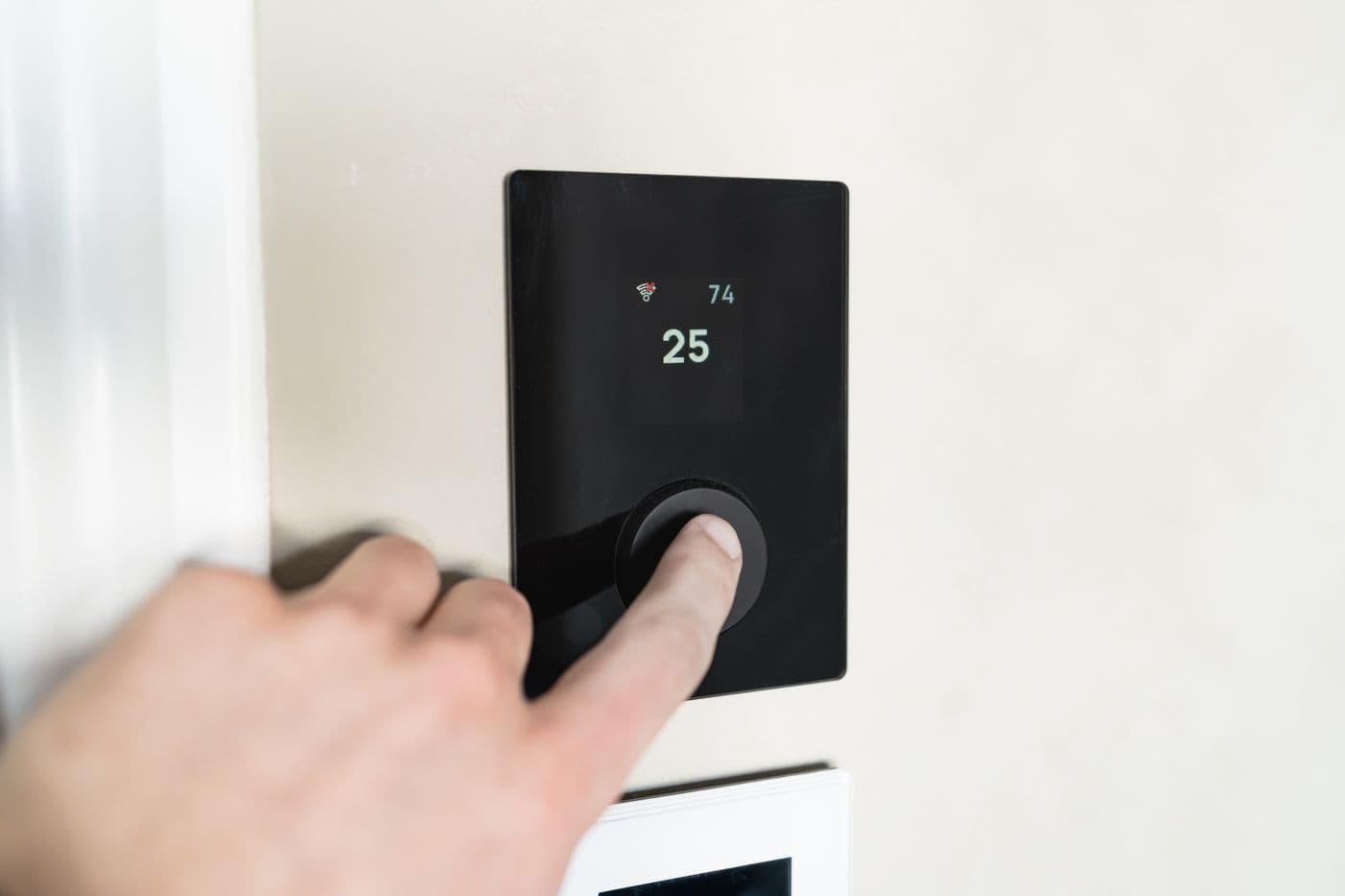 A hand pressing the dial on a black wall-mounted smart thermostat showing the temperature