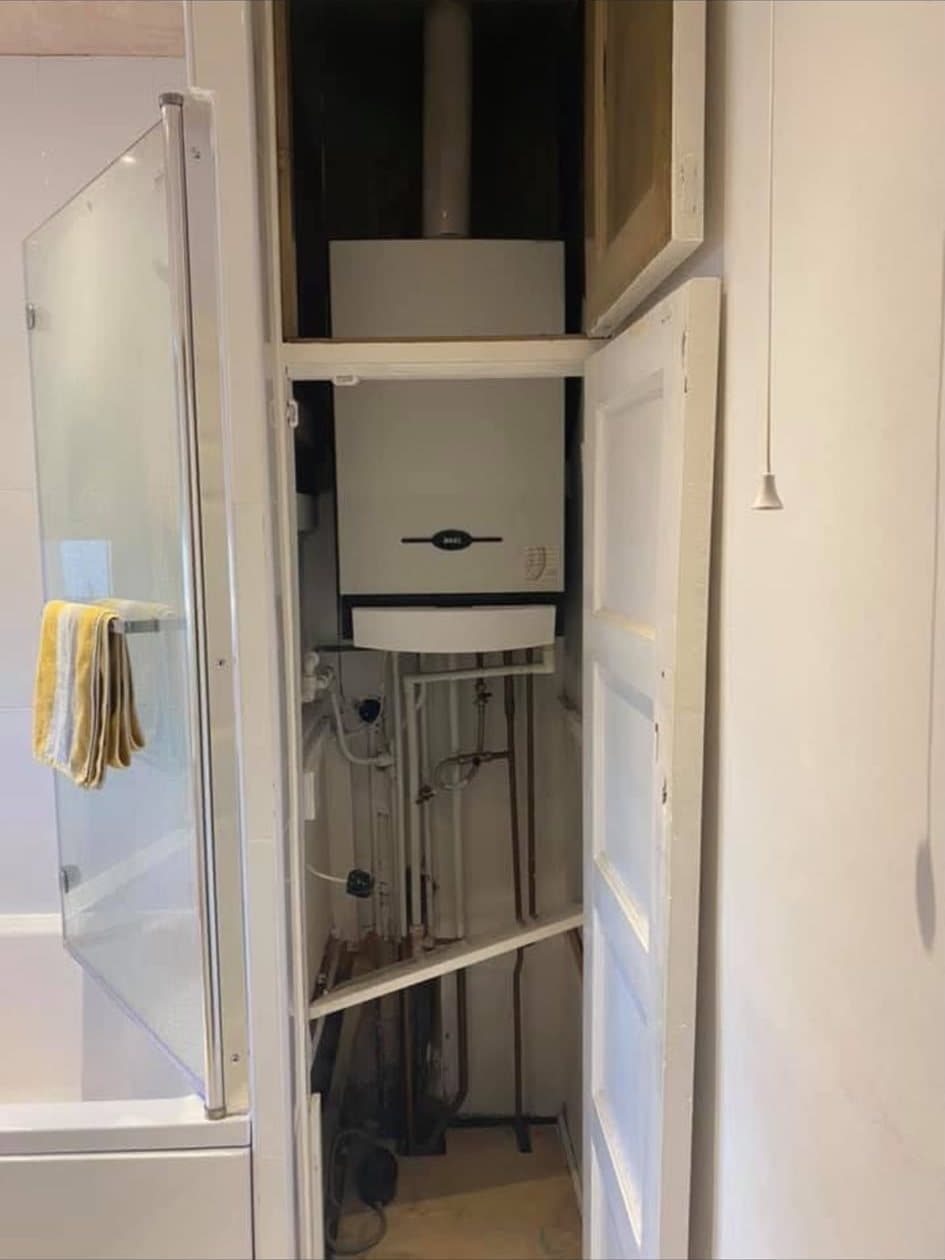 A boiler fitted in an airing cupboard off a bathroom, its flue rising above it and pipework dropping below