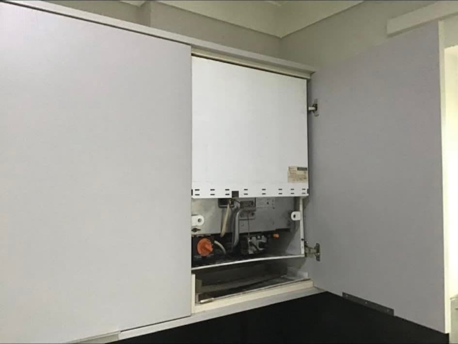 A boiler inside a kitchen cupboard with its front cover removed, showing the controls and connections
