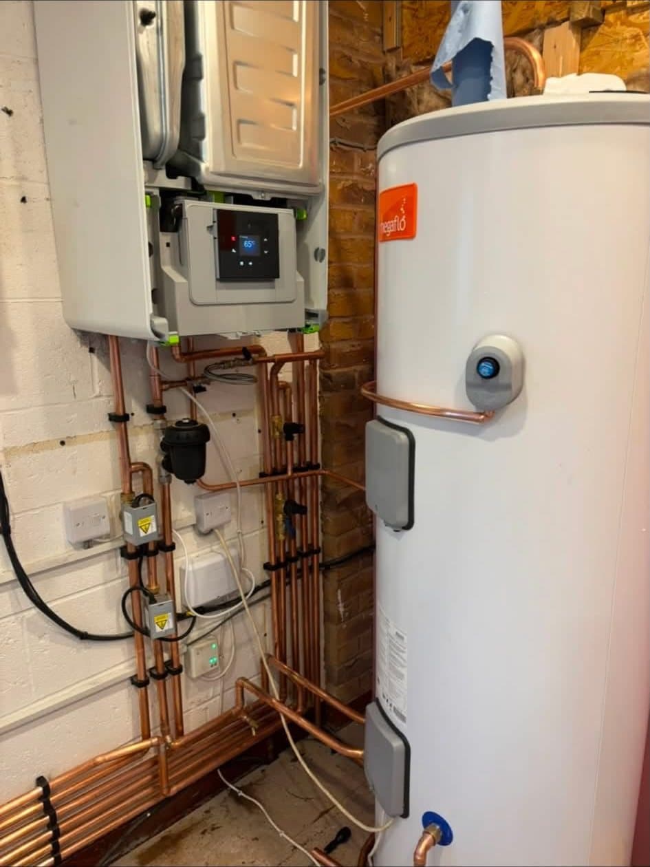 A boiler with its front cover off beside a white unvented hot water cylinder, joined by neat runs of copper pipework, valves and wiring