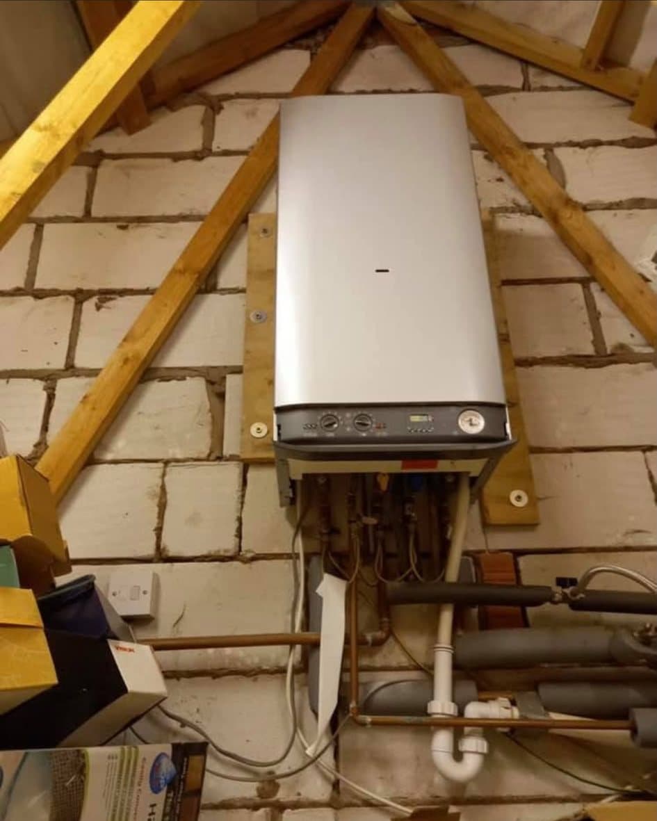 The old boiler on a blockwork wall in a loft, with loose cables and bare pipework hanging beneath it